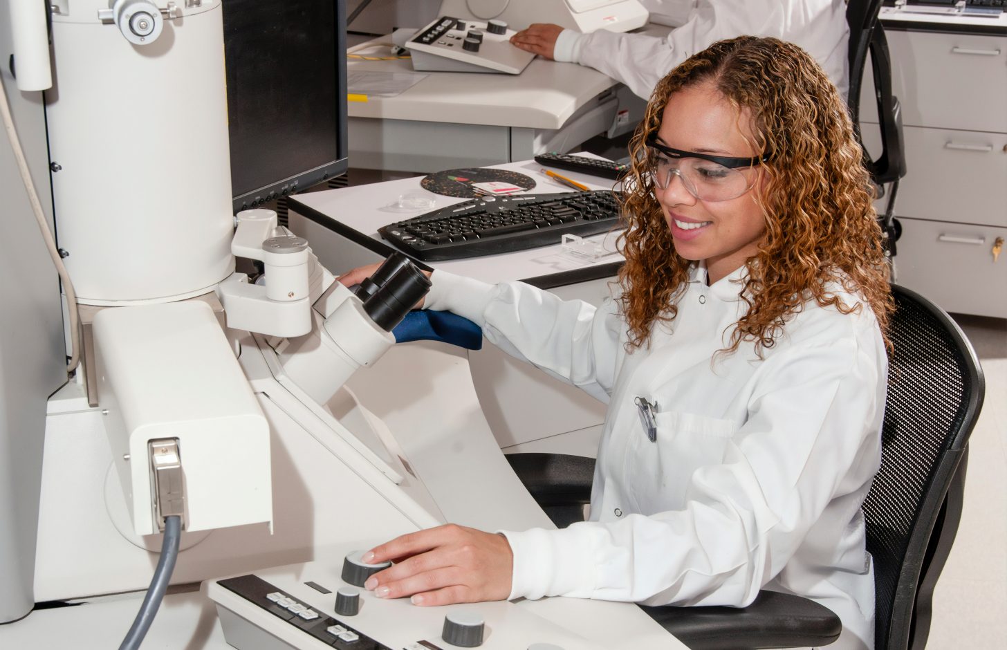 A young black woman in lab coat and goggles experiments on lab equipment