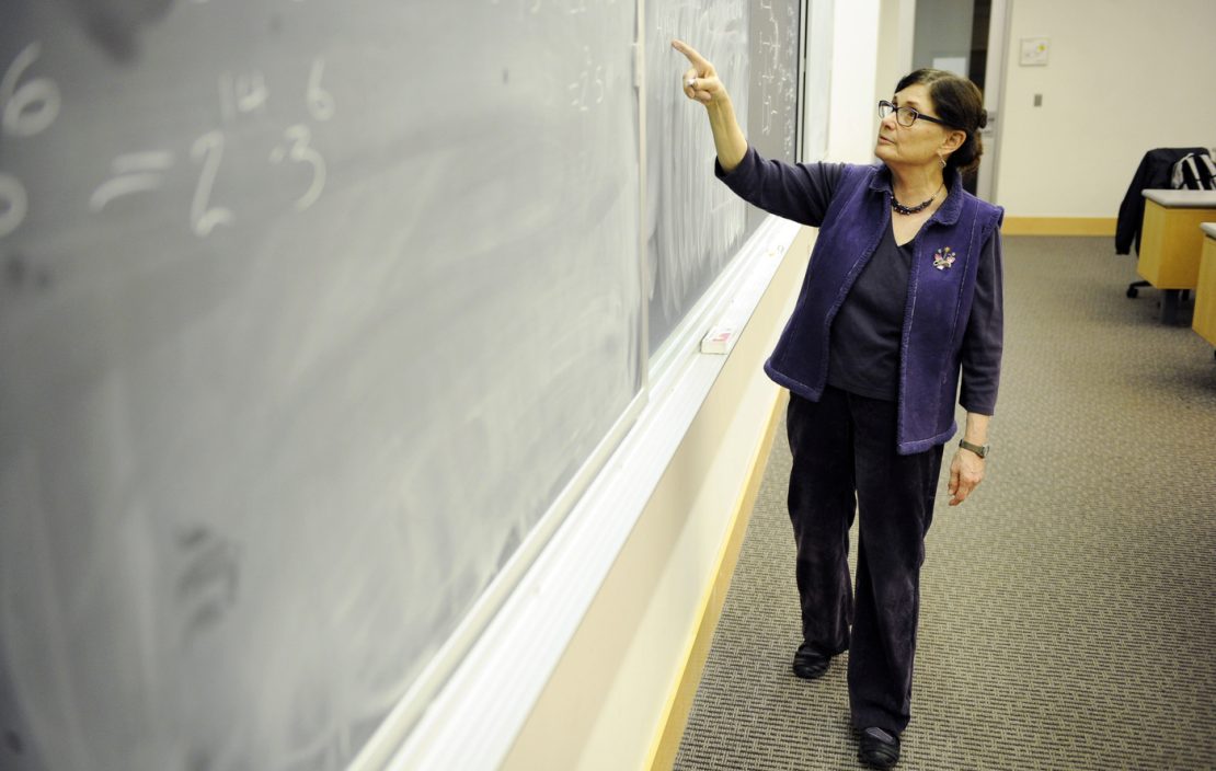 A woman gives a lecture to her students, gesturing at a blackboard.